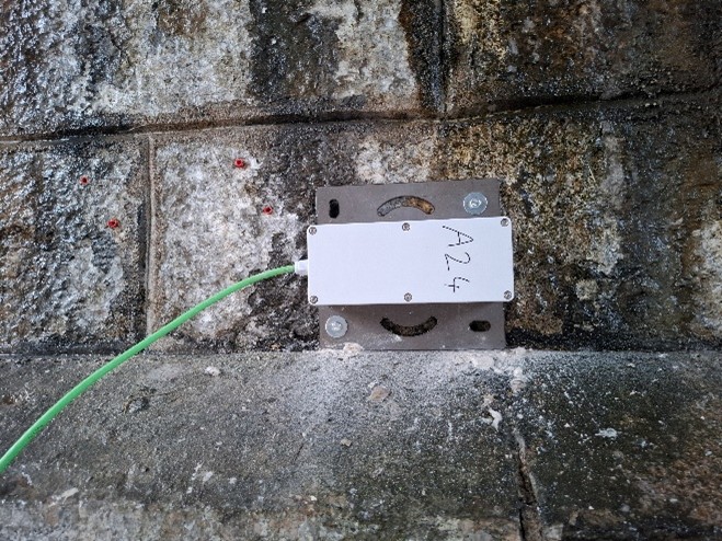 Boîtier de capteur de surveillance fixé sur un mur en pierre, relié par un câble vert, utilisé pour le suivi structurel d’un ouvrage